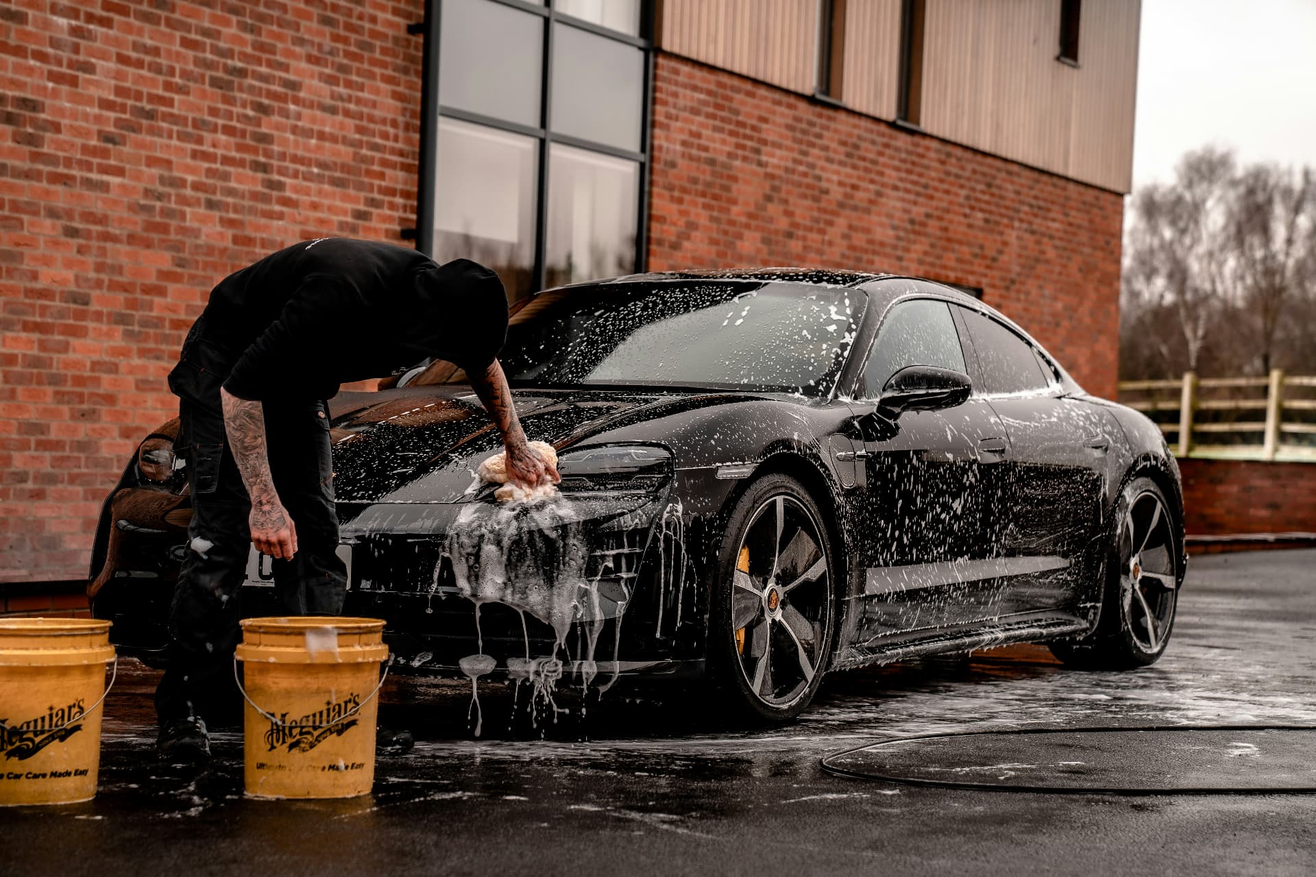 Detailer washing a black sports car with soap suds in front of a brick building